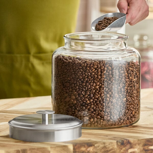 A large clear glass jar filled with coffee beans, shown with a brushed metal lid resting beside it.