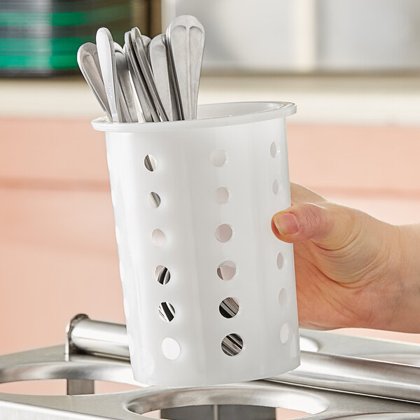 A hand holding a white Vollrath flatware cylinder filled with silver spoons.