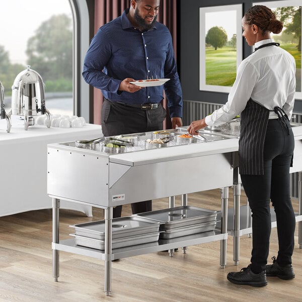 A man and a woman standing at a ServIt cold food table with metal containers.