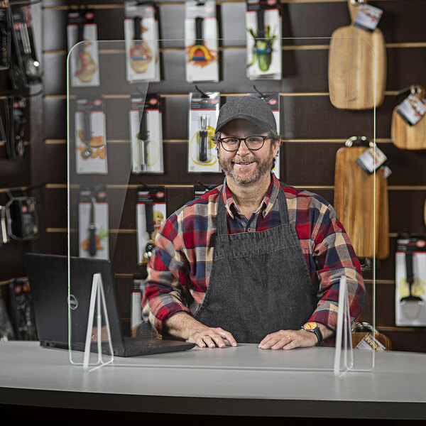 A man in a hat and apron behind a Tablecraft clear acrylic countertop safety shield.