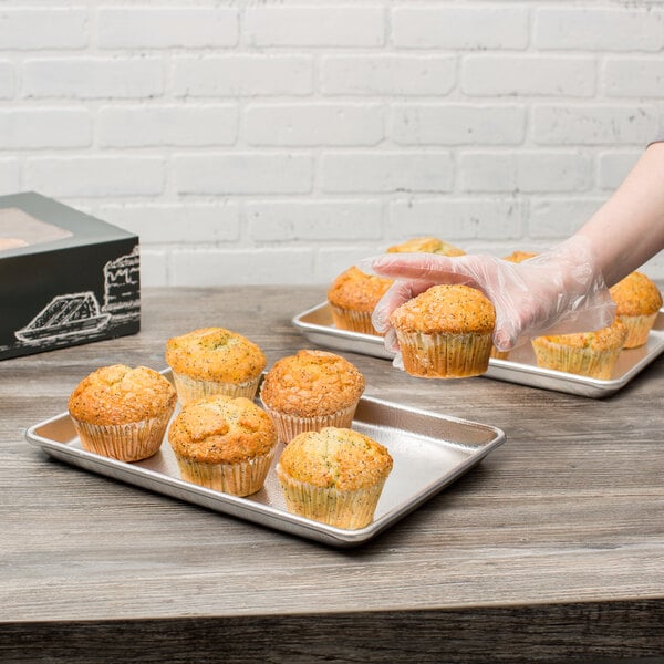 A person wearing gloves holding a Chicago Metallic bakery display tray with a muffin inside.