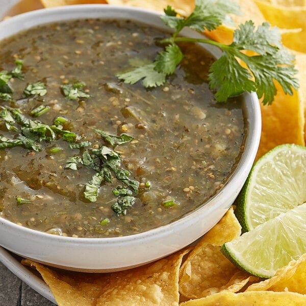 A bowl of green sauce with chips and limes on a table.