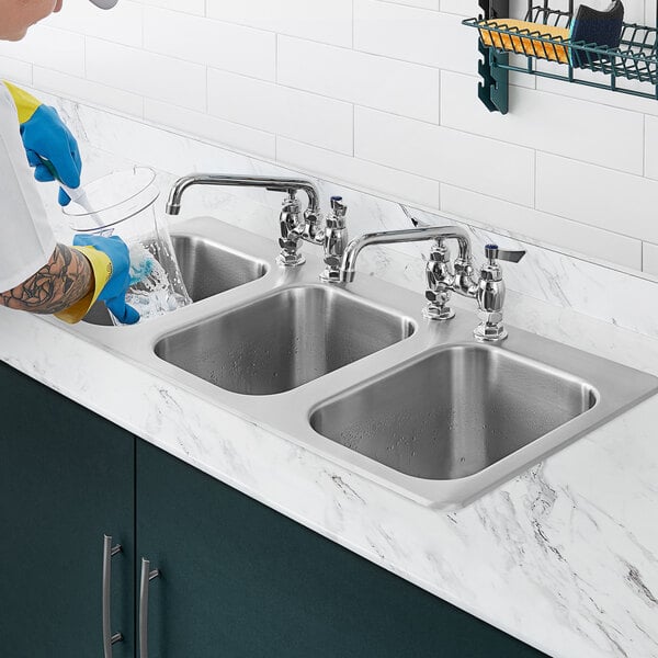 A person washing dishes in a Waterloo stainless steel three compartment drop-in sink with a swing faucet.