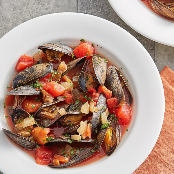 A white plate with a bowl of soup with mussels and tomatoes on it.