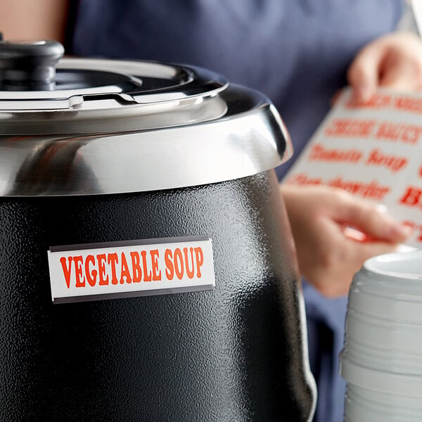 A woman using a silver Avantco magnetic label on a white container of vegetable soup.