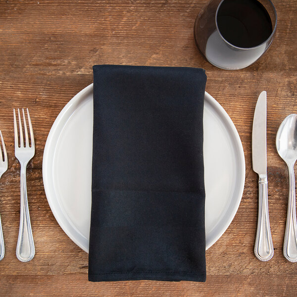 A black Milan satin band cloth napkin folded on a plate with silverware on a table.