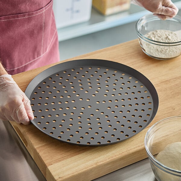 A person using an American Metalcraft hard coat anodized aluminum perforated pizza pan to make pizza.