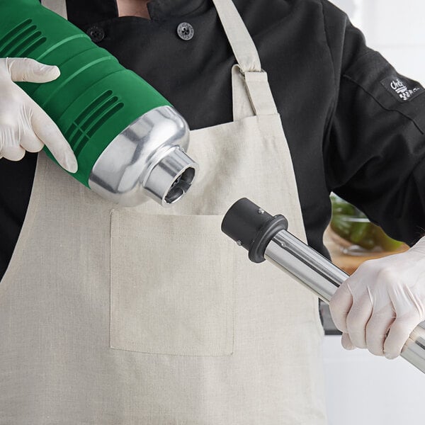 A person in a white apron and gloves using an AvaMix heavy-duty immersion blender with a metal tube attachment.
