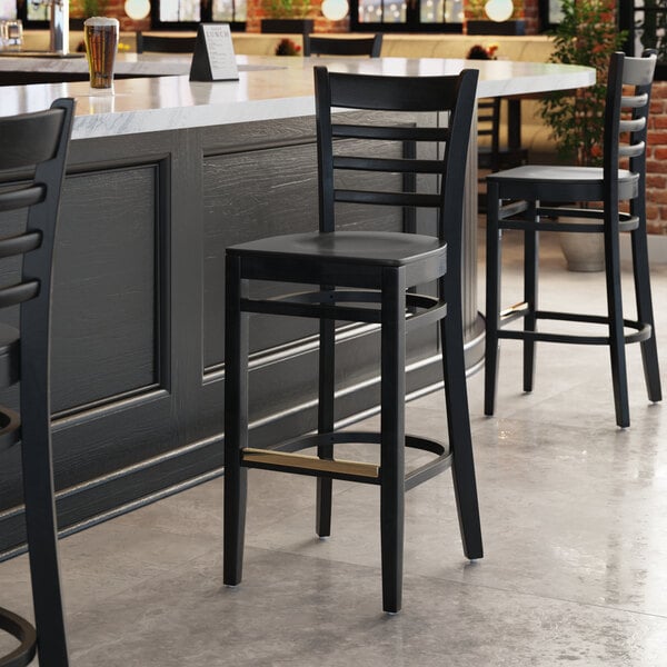 A Lancaster Table & Seating black wood ladder back bar stool with a black wood seat next to a table in a bar.