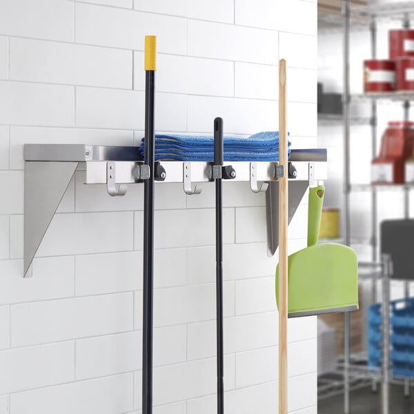 A Regency stainless steel utility shelf with mop and broom holders holding mops and brooms.