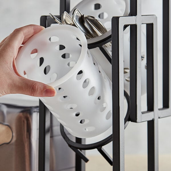 A hand holding a white Tablecraft polypropylene container with holes filled with spoons.