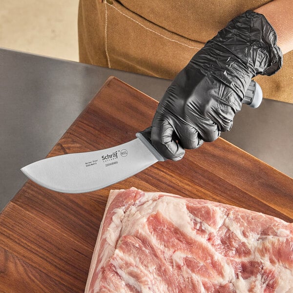 A person in black gloves using a Schraf skinning knife to cut meat on a wooden surface.