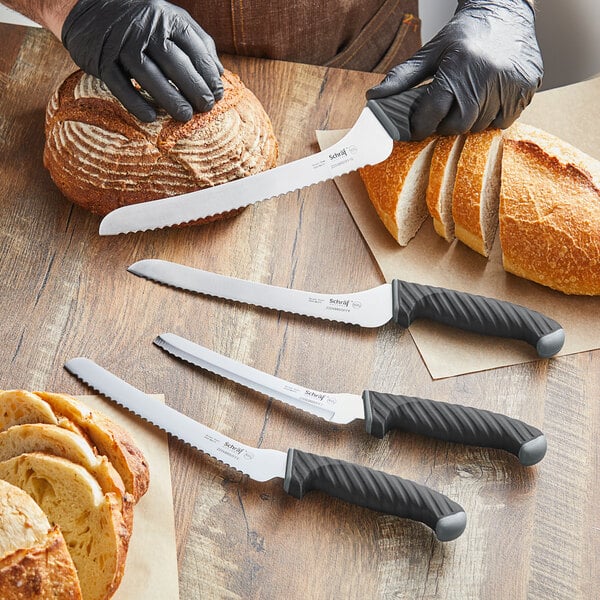 A hand in a black glove uses a Schraf serrated bread knife with a white handle to cut a loaf of bread.