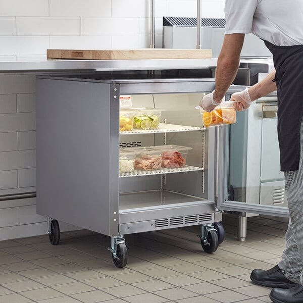 A man in a white apron putting a container of food into a Beverage-Air undercounter refrigerator.