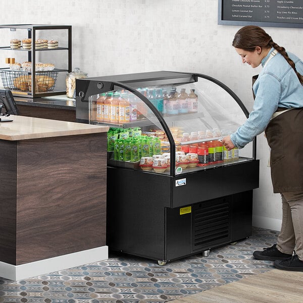 A woman looking at an Avantco black horizontal air curtain merchandiser on a hospital cafeteria counter.