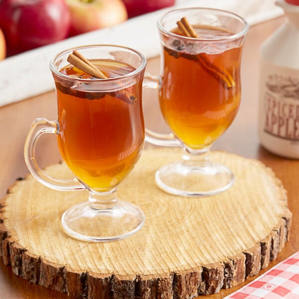 Two glasses of brown Mountain Cider Company spiced apple cider with cinnamon sticks on a wood surface.