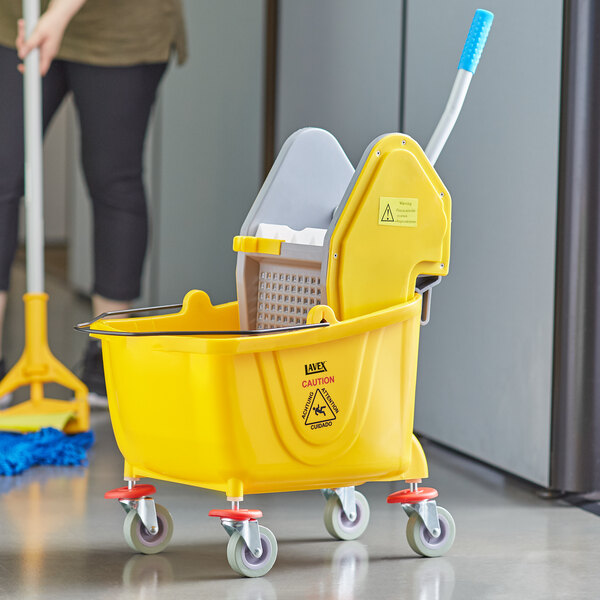 A woman using a yellow Lavex mop bucket with a down press wringer to clean a mop.