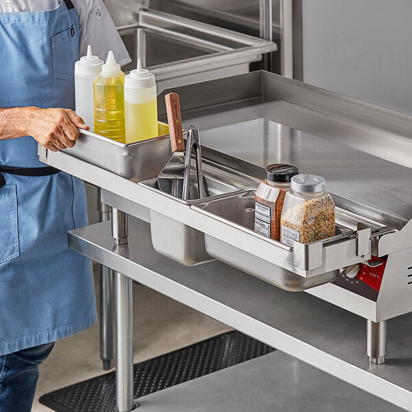 A man in a blue apron standing next to a Universal 36" Griddle Condiment Rail filled with metal containers.