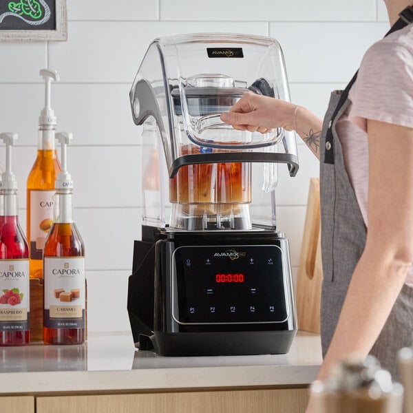 A commercial blender with a sound enclosure and digital touchpad, shown on a countertop next to flavored syrups.