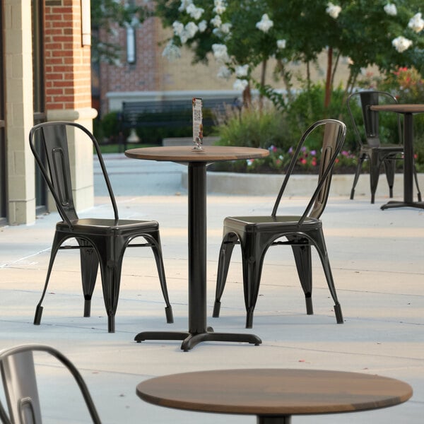 A table with a Lancaster Table & Seating Excalibur round table top with a textured farmhouse finish and black chairs on a patio.