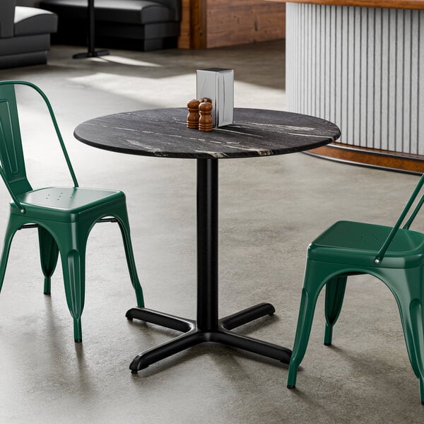 A round black table with a smooth finish, supported by a central pedestal base, shown with two green metal chairs.