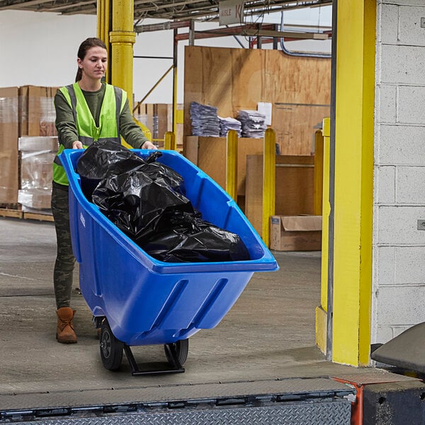 A woman in a safety vest pushing a blue Lavex tilt truck full of black bags.