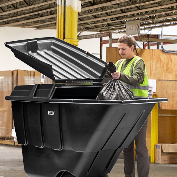 A woman in a safety vest putting a garbage bag into a large black Lavex tilt truck lid.