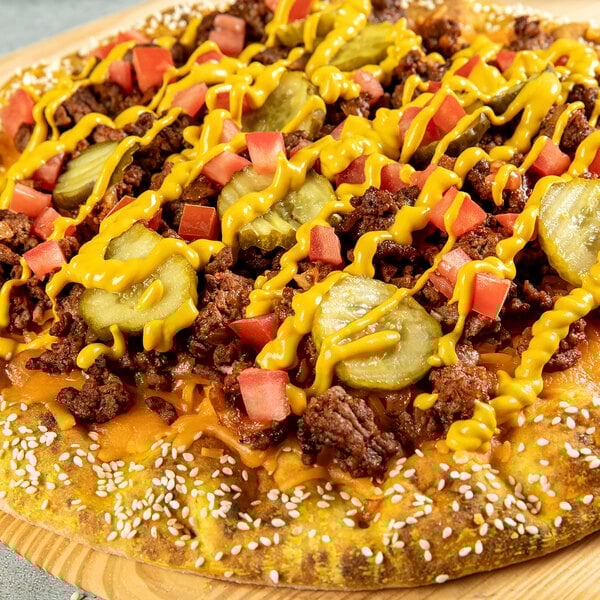A cheeseburger-inspired pizza topped with ground beef, pickles, diced tomatoes, cheddar cheese, and drizzled with yellow mustard on a sesame seed crust.