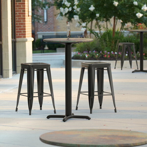 A Lancaster Table & Seating round counter height table with a textured metal finish and two stools on a patio.
