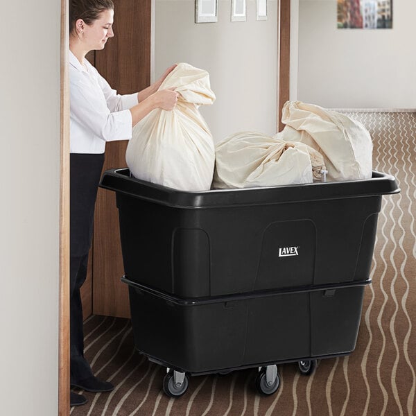 A woman pushing a large white bag into a black plastic bin on wheels.