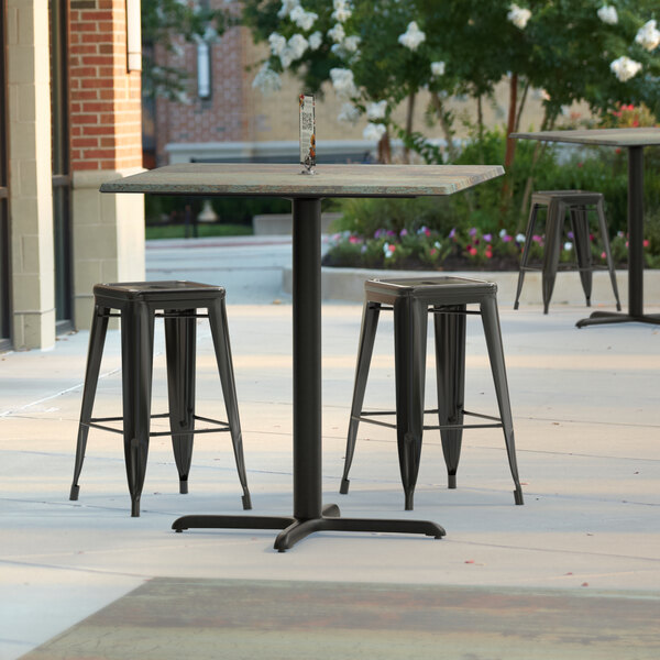 A Lancaster Table and Seating counter height table with a textured metal finish and cross base plate on a patio with three stools.