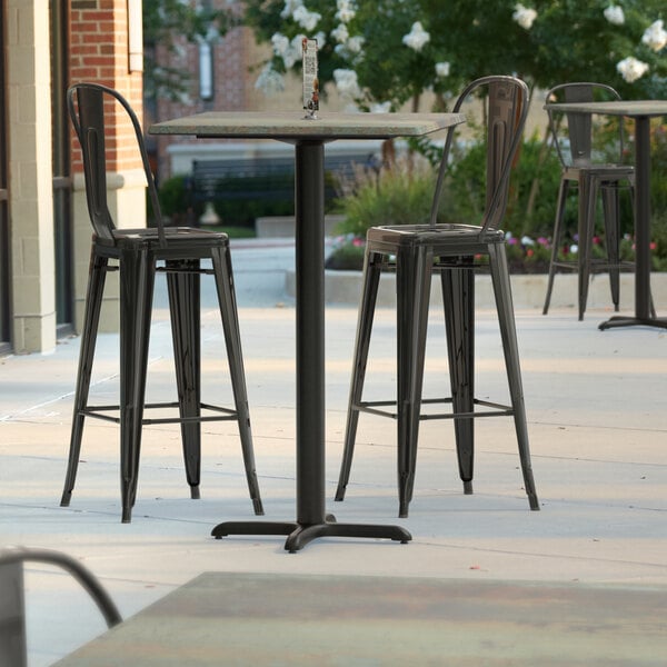 A Lancaster Table & Seating bar height table with a textured canyon metal finish and cross base plate on a patio with chairs.