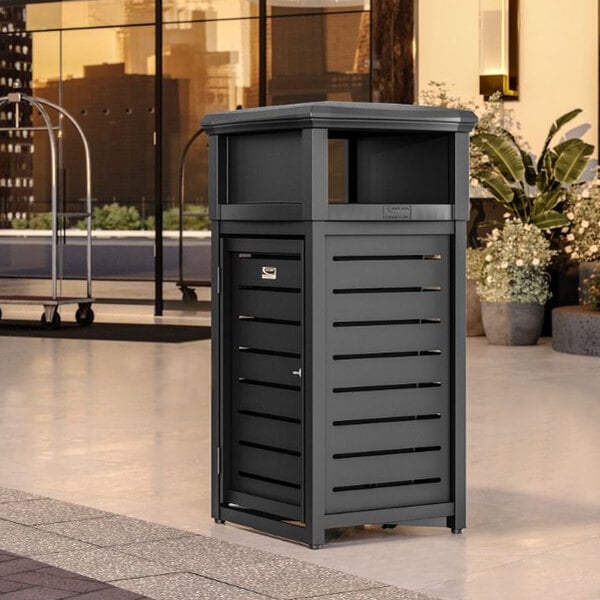 A black Suncast square metal trash can with the lid open outside on a sidewalk.