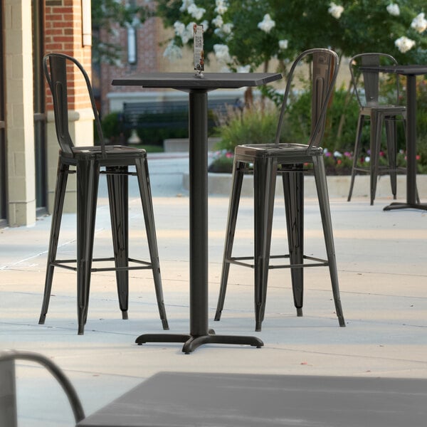 A Lancaster Table & Seating bar height table with a cross base on an outdoor patio with three black chairs.