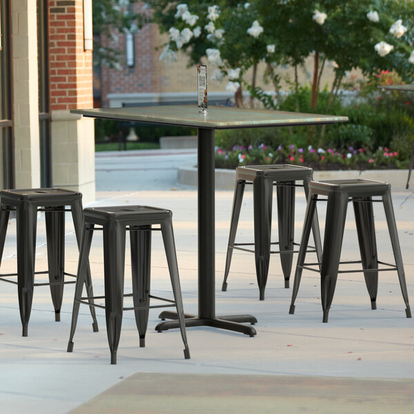 A Lancaster Table & Seating rectangular counter height table with a textured metal finish and cross base plate on an outdoor patio with four black stools.