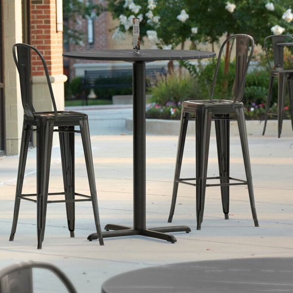 A Lancaster Table & Seating bar height table with a cross base on a patio with three black metal chairs.