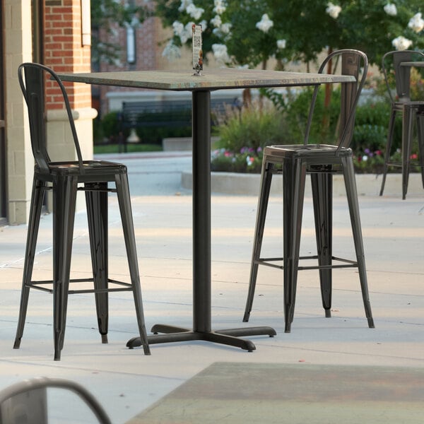 A Lancaster Table & Seating bar height table with a textured metal finish and cross base plate on a patio with black bar stools.