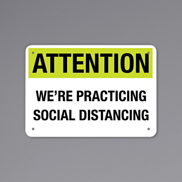 A black and yellow rectangular decal on a wall that says "Attention / We're Practicing Social Distancing" with black text.