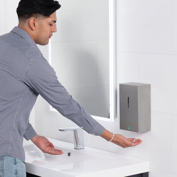 A person is using a wall-mounted, stainless steel automatic soap dispenser above a white sink in a modern restroom.