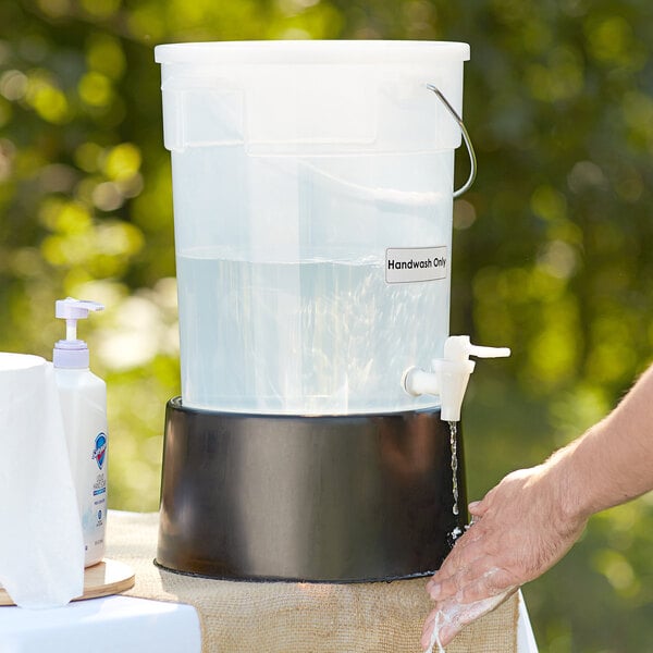 A person washing their hands in a white round beverage dispenser with a black base.