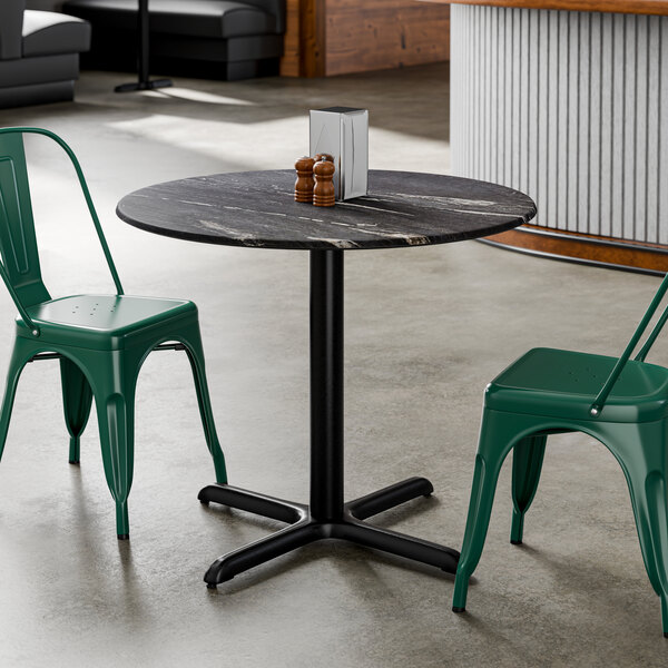 A round black table with a smooth finish and cross base plate, accompanied by two green metal chairs.