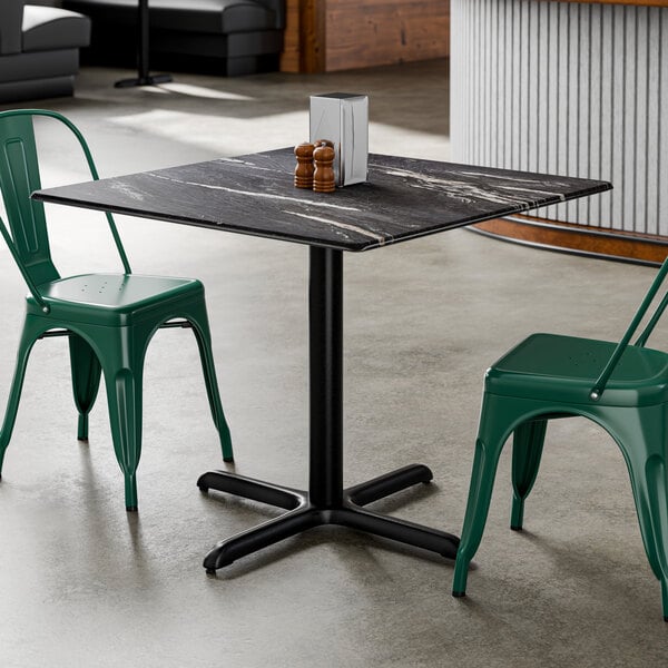 A square standard height table with a smooth dark finish and a black cross base plate, accompanied by two green metal chairs.