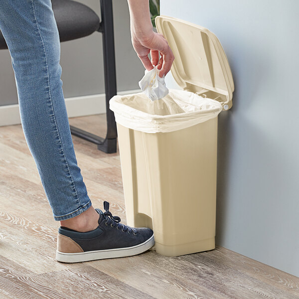 A woman putting a white bag in a Lavex beige rectangular step-on trash can.