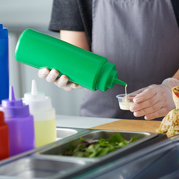 A person pouring sauce from a green Choice Wide Mouth Squeeze Bottle into a small cup.
