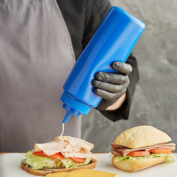 A person in black gloves and apron holding a Choice blue wide mouth squeeze bottle over a sandwich with meat and vegetables.