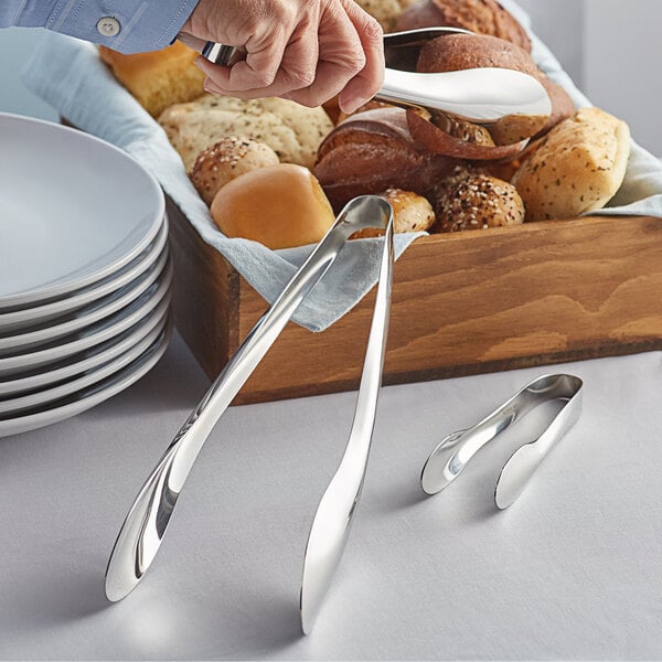 A person using Acopa stainless steel tongs to grab bread from a basket.