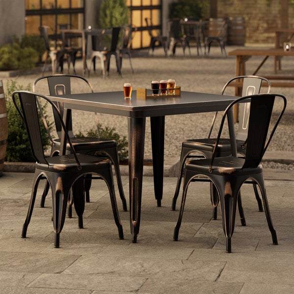 A Lancaster Table & Seating distressed copper table with a black metal base and four chairs outside.