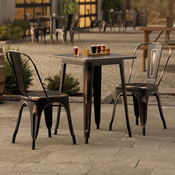 A Lancaster Table & Seating distressed copper table with two chairs on an outdoor patio.