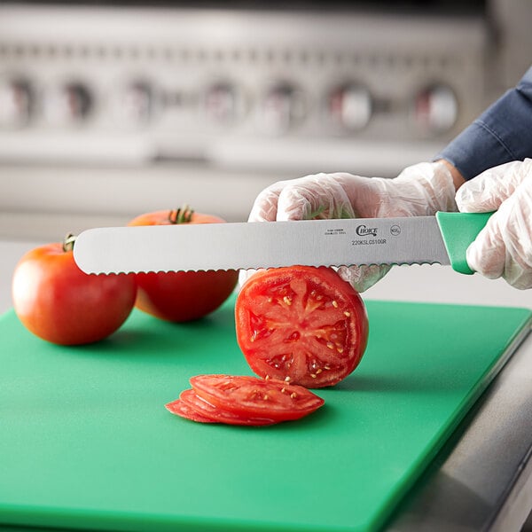 A person using a Choice serrated bread knife with a green handle to cut a tomato on a white rectangular object.