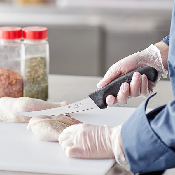 A person in a white coat using a Choice 6" Curved Stiff Boning Knife to cut chicken on a counter in a professional kitchen.